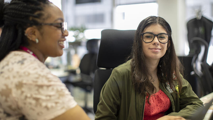 A supported internship learner sitting at a desk by a computer. She is being supported with her work by another woman.