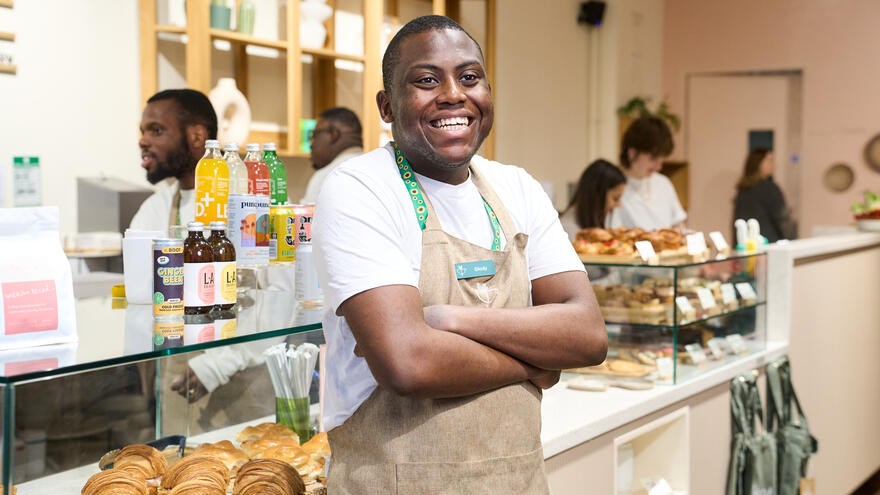 A man standing by a counter in a cafe. He is wearing an apron and smiling at the camera with his arms folded.