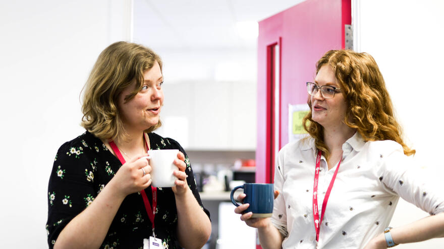 Two supported internship tutors stand by a door talking to each other. They are holding cups of tea.