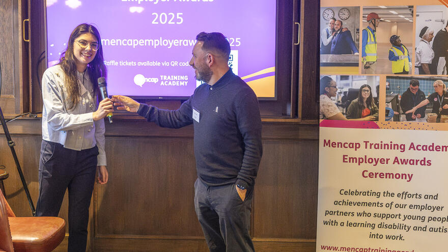 Mencap colleagues, Natalie Duo and Mark Capper presenting with a microphone in front of a screen at the 2025 Mencap Training Academy Employer Awards.