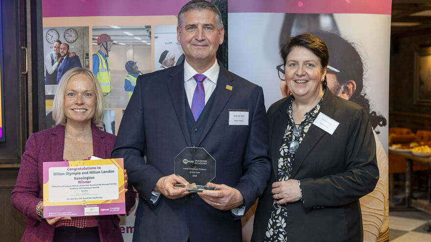 Three people standing and smiling at the camera. The woman on the left is holding a certificate and the man in the middle is holding a glass trophy.