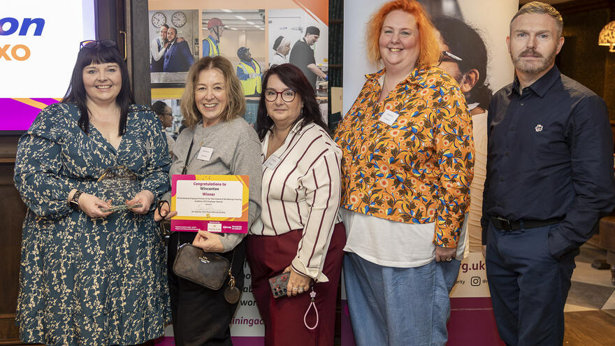 5 people standing and smiling at the camera. The woman on the left is holding a glass trophy. The woman on her right is holding up a certificate.